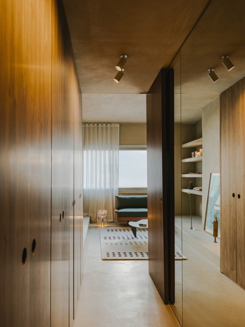 Modern hallway with wooden closets, mirrored wall, and a view into a sitting area with a window, couch, shelves, and a patterned rug.