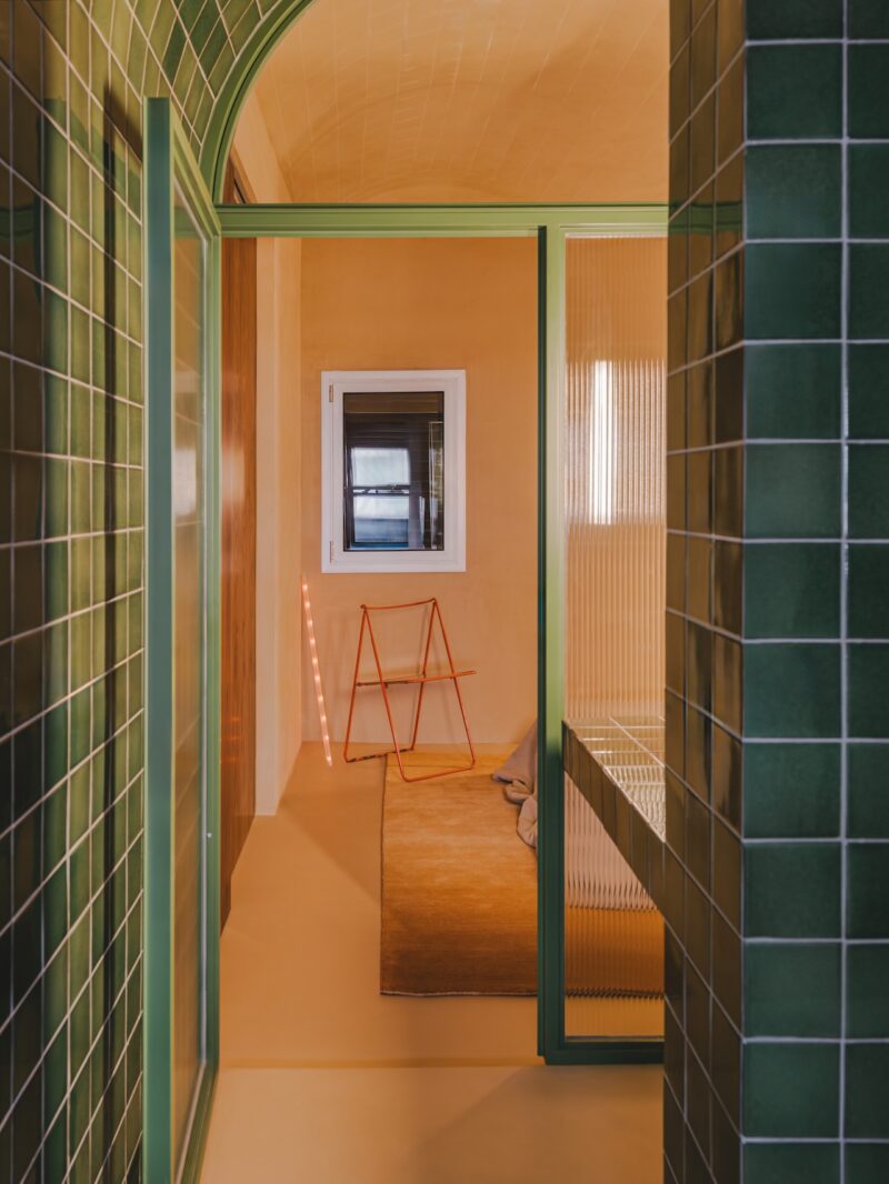 A green-tiled doorway leads to a room with orange walls, a window, a tan rug, a pink ladder, and a cushion on the floor. Vertical light strip glows by the wall.