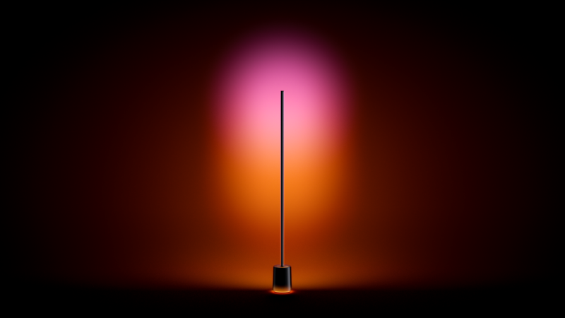 A tall, thin floor lamp emits a gradient light with pink and orange tones, creating a soft glow against a dark background and enhancing the ambience with layered lighting.