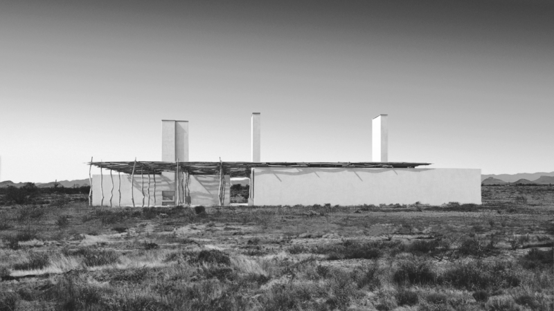 Rectangular modern building with three tall chimneys and a flat roof stands in an open, arid landscape with mountains in the distance under a clear sky.