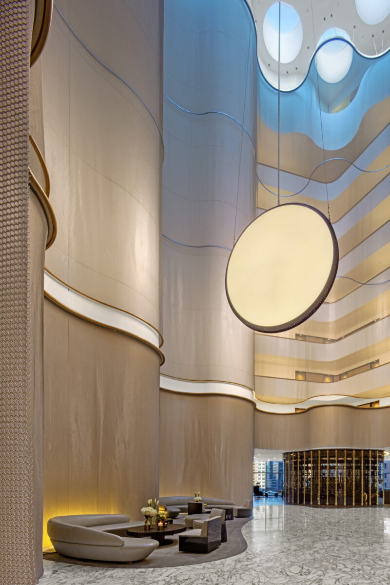 Spacious modern hotel lobby with tall curved beige walls, a large circular light fixture, marble floors, and contemporary seating arrangements.