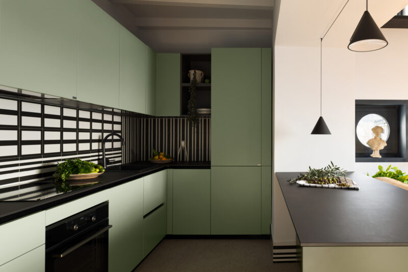 Modern kitchen with matte green cabinets, black countertops, striped black and white backsplash, and pendant lights. The aesthetic captures the contemporary essence of La Casa Olive e Capperi. A bust sculpture is visible through a window in the background.
