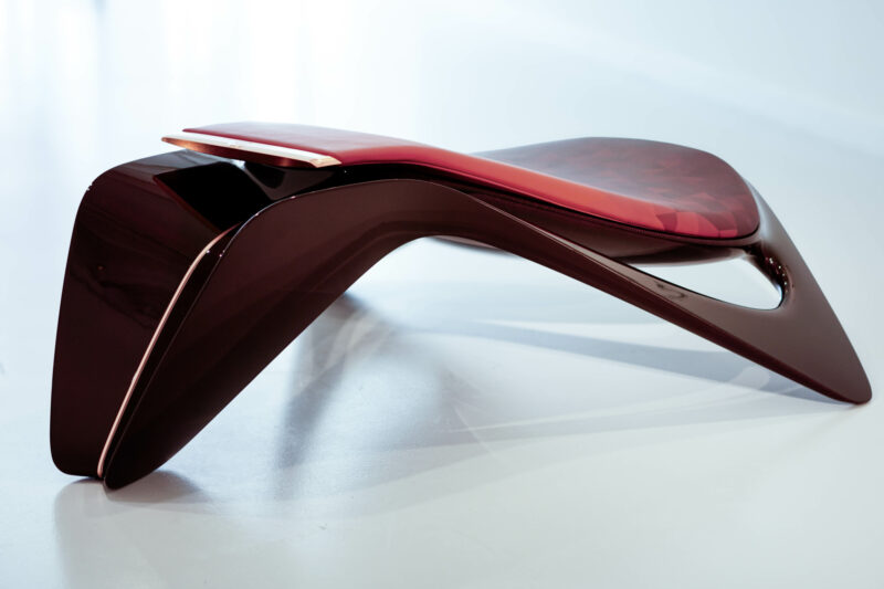 A modern, sculptural chaise longue from the Ennua Element 01 collection features a sleek, curved design in dark red and black, set against a plain white background.