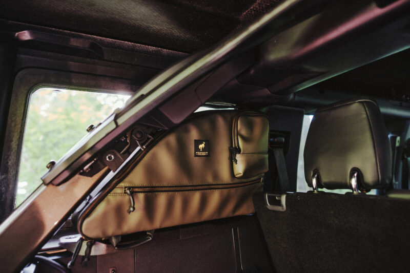 Tan storage bag with a black logo is secured behind the front seats of a vehicle, attached to the rear side of the driver’s seat.