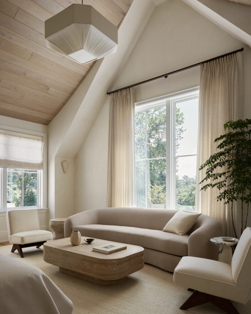 Neutral-toned living room with a curved sofa, wooden coffee table, two chairs, large windows, and a potted plant. Light streams in, highlighting minimalist decor and natural materials.