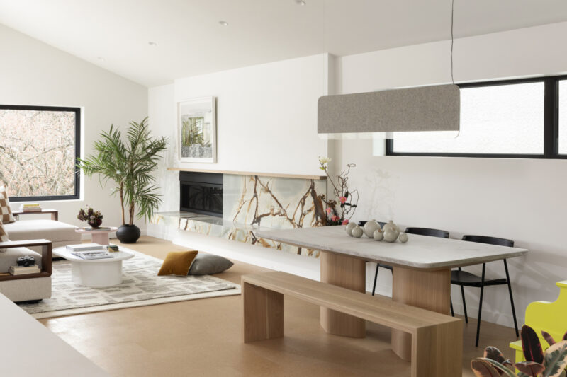 Modern living-dining room with neutral tones, marble fireplace, large windows, green plant, light wood furniture, and minimalist decor.