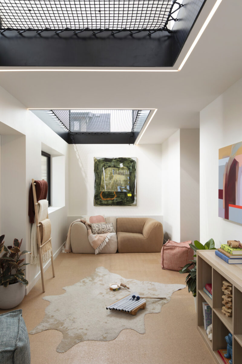 Modern living room with beige furniture, a cowhide rug, bookshelves, and abstract art. A skylight with a netted platform allows natural light into the bright space.