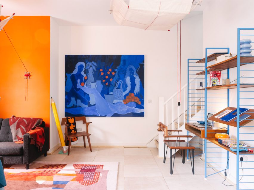 Photo of the main room of the UAL Atterbury Flat, with white walls and bright decoration including a big blue painting, an oversized pencil sculpture, a minimal wooden chair, and a study nook with a vast canvas pendant light overhead