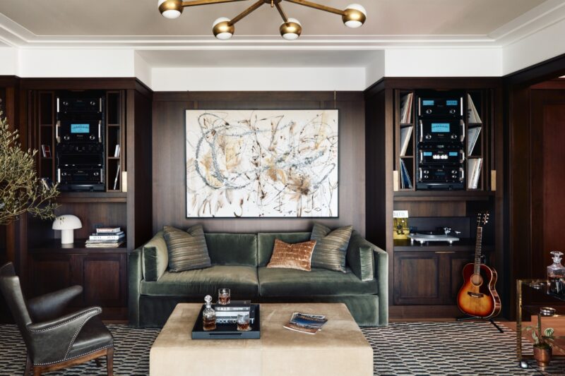 A living room with a green velvet sofa, abstract wall art, audio equipment shelves, a guitar, a black chair, a beige ottoman, and a mid-century chandelier.