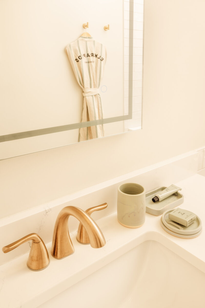A bathroom sink with gold faucet, soap dish, toothbrush holder, and a robe hanging on the wall reflected in the mirror at Dexter Hotel Michigan.