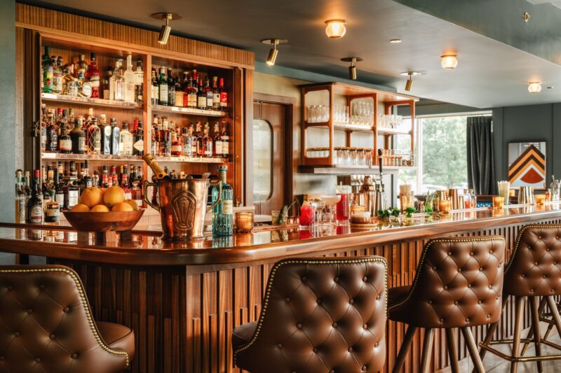 A stylish bar with a wooden counter, leather bar stools, and shelves stocked with bottles, glasses, and fruit. Warm lighting creates a cozy atmosphere.