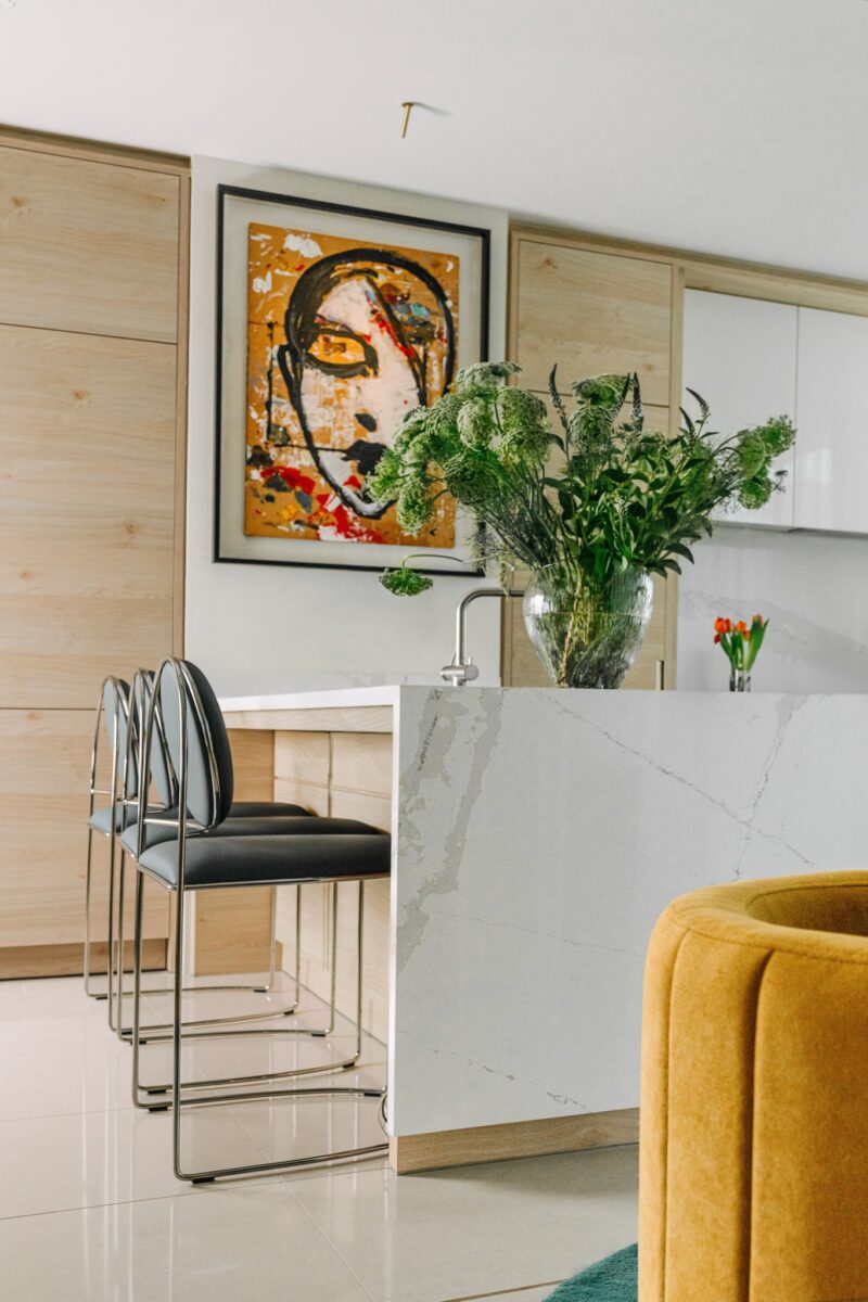Modern kitchen with a marble island, black chairs, a large vase of greenery, abstract face artwork on the wall, and light wood cabinetry.