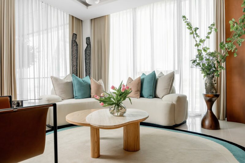 Modern living room with a curved white sofa, assorted pillows, wooden coffee table with pink tulips, large windows with sheer curtains, and plants on the side.