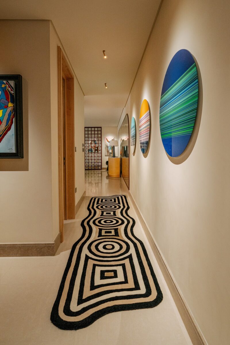 A hallway with a patterned black and white rug, abstract circular art on the walls, and neutral-toned decor leading to a distant room.