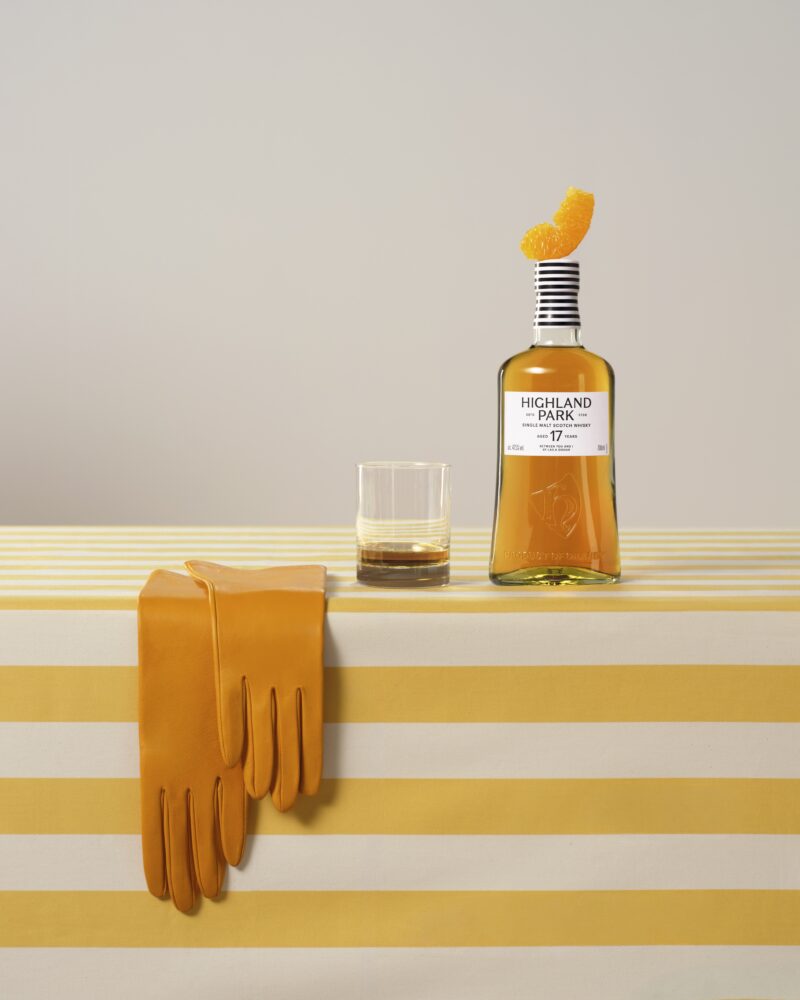 A bottle of Highland Park 17-Year-Old whisky, a glass with whisky, and a pair of yellow gloves are arranged on a yellow and white striped tablecloth.