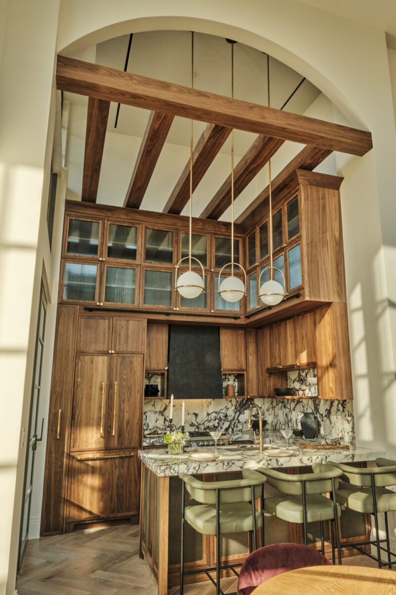 Modern kitchen with wooden cabinets, marble countertops and backsplash, three pendant lights, green barstools, and high ceilings with exposed beams.