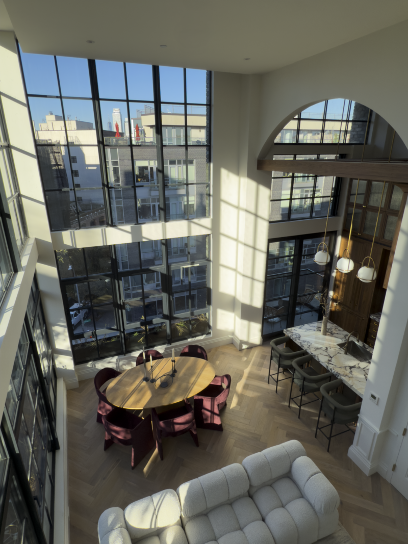 Modern loft with large floor-to-ceiling windows, a round dining table with red chairs, a marble kitchen island with stools, and a white sofa in an open, sunlit space.