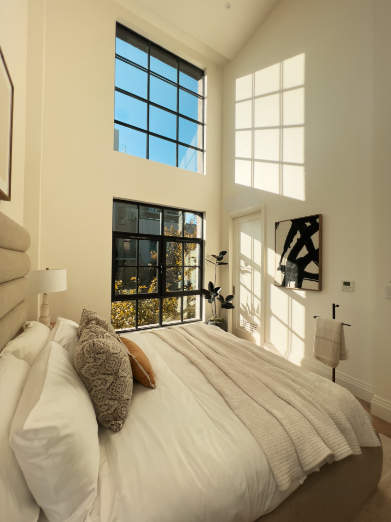 A sunlit bedroom with tall windows, a made bed with white and beige bedding, a plant, framed art on the wall, and shadows cast on the walls and floor.