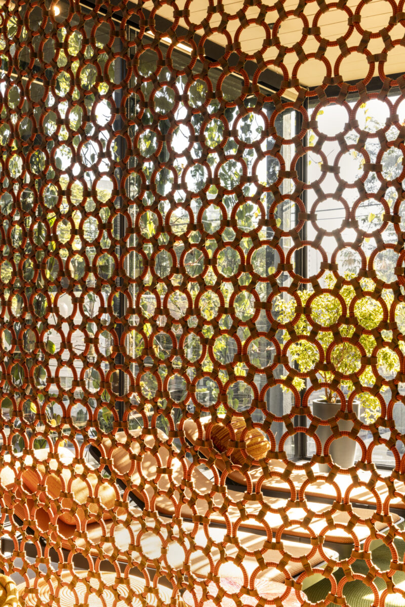 Orange circular lattice screen in front of large glass windows, with sunlight streaming through and trees visible outside. Modern furniture is partially visible behind the screen.
