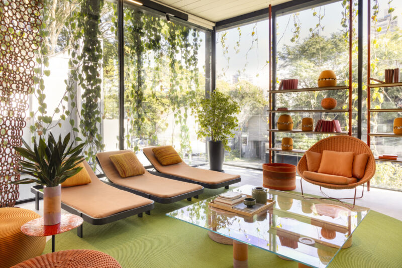 Modern sunroom with orange lounge chairs, circular chair, glass coffee table, potted plants, and floor-to-ceiling windows covered in hanging greenery. Shelving holds orange decor items.