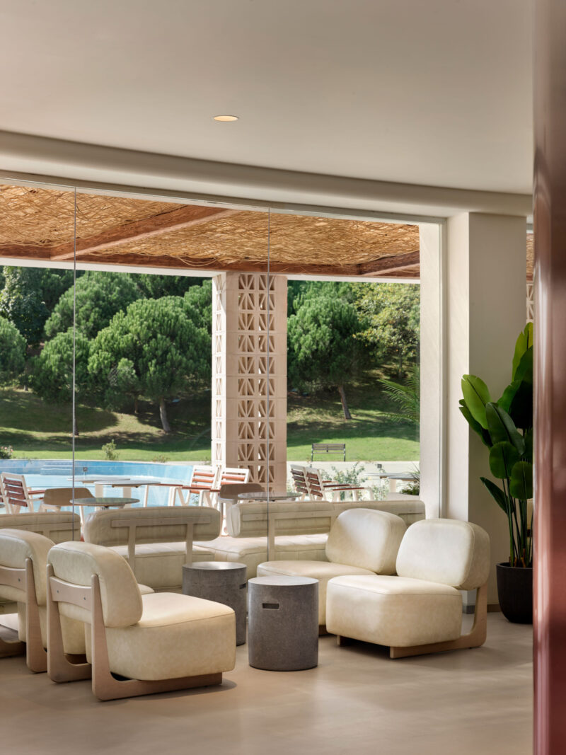 Modern Local People cafe lounge area with light-colored chairs and stone tables, looking out through floor-to-ceiling glass at a swimming pool and trees.