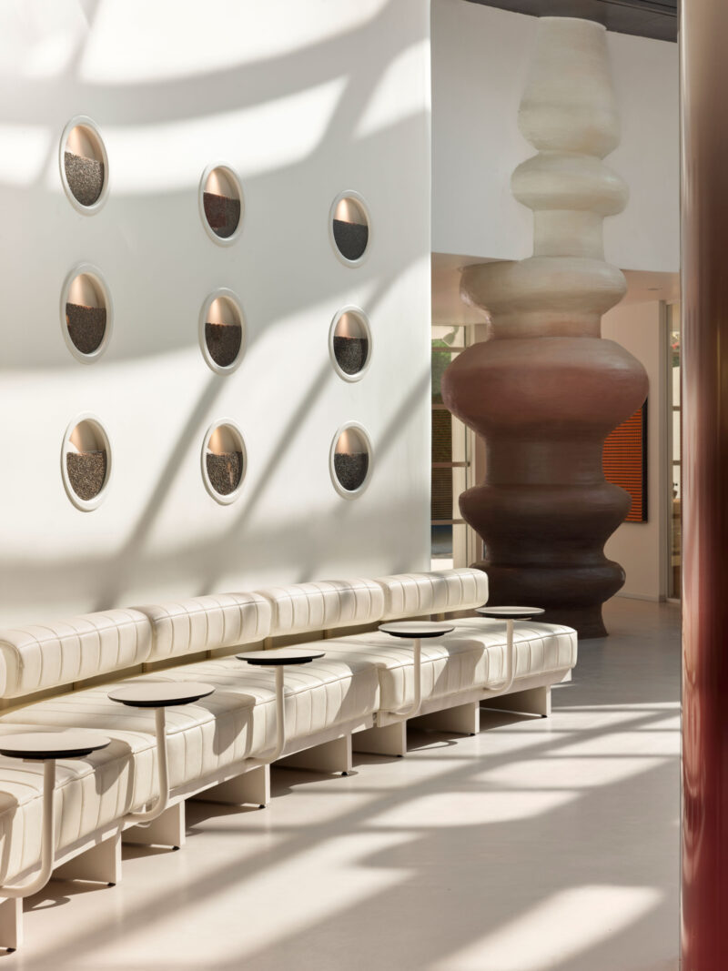 Long white modular bench with attached small circular tables, set against a wall featuring nine circular insets filled with dark grains and a large abstract sculptural column.