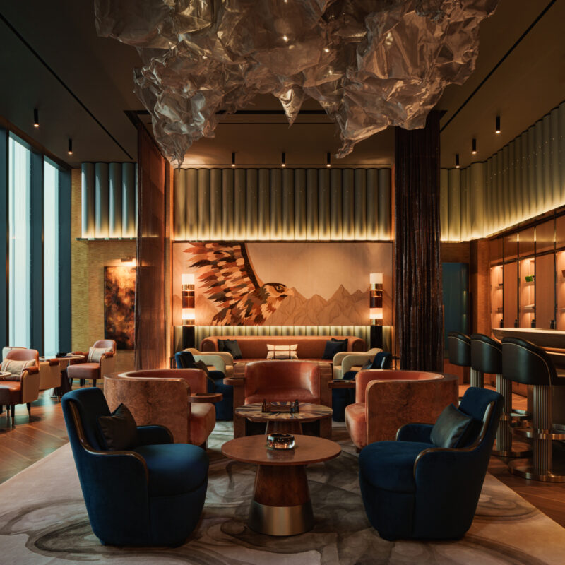 A lounge area featuring blue and orange velvet armchairs around circular wooden tables, a mural of a falcon on the back wall, and a large sculptural light fixture hanging from the ceiling.