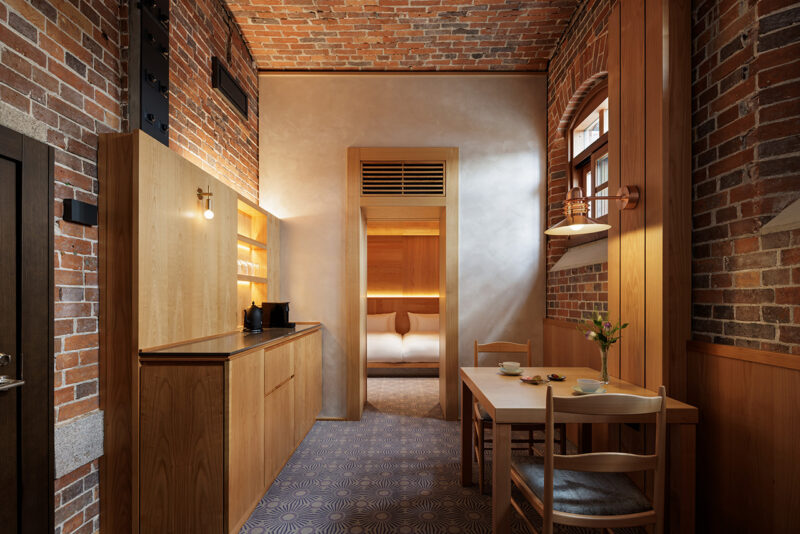 A hotel room with exposed brick walls, a wooden kitchenette, and a dining table for two, with a view into a bedroom featuring a bed with white linens.