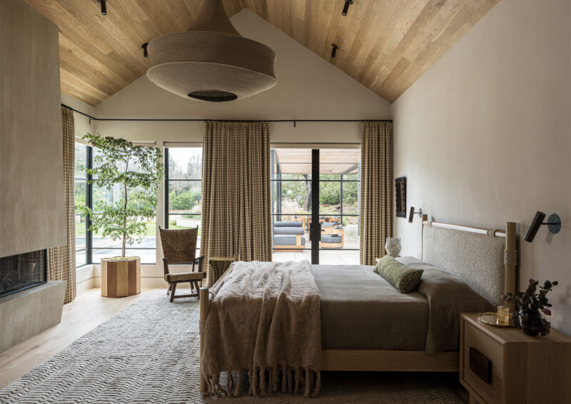 A bedroom with a vaulted wooden ceiling, a bed with a textured headboard, a fireplace, and glass doors opening to a patio.