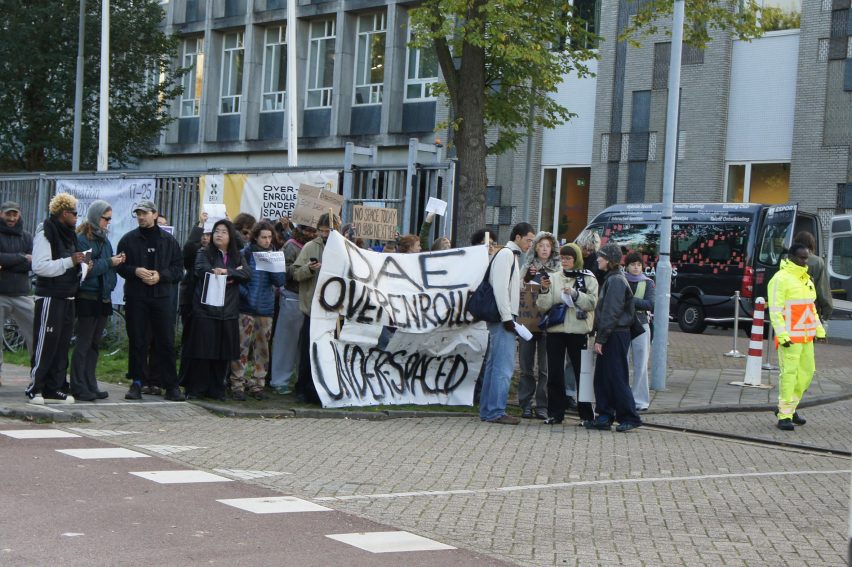 Photo of a huddle of students protesting at the gates to the Design Academy Eindhoven building, holding a banner that reads DAE overenrolled under-spaced