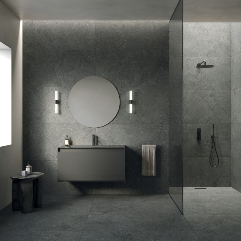 A modern bathroom with grey stone walls, a floating vanity with a circular mirror, and a walk-in shower.