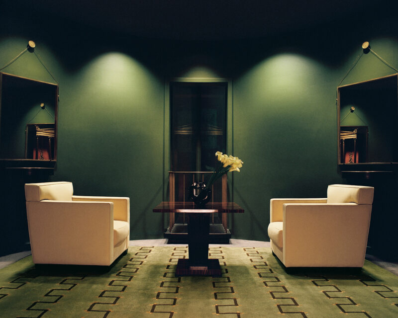 Two white armchairs face each other across a dark wood table with a vase of flowers, set on a patterned green rug against a green wall with mirrors and accent lighting.