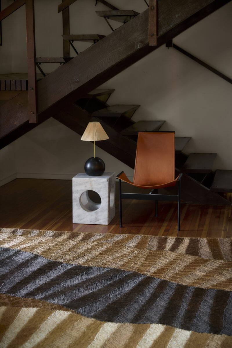 A brown leather chair, marble side table with a black lamp, and a patterned rug sit beneath a wooden staircase in a modern interior.