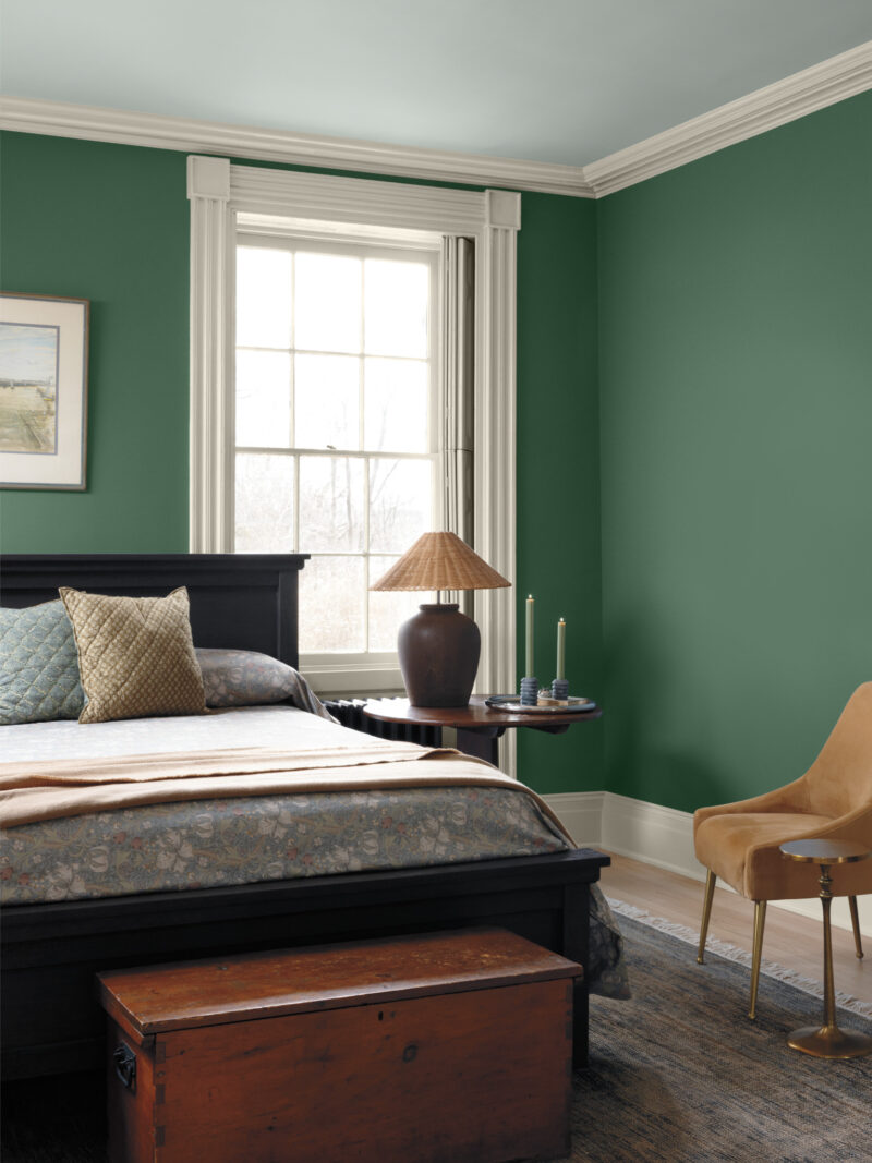A bedroom with Benjamin Moore Color of the Year 2027 dark green walls, a black bed frame with a patterned quilt, a wooden chest, and a tan chair beside a window.