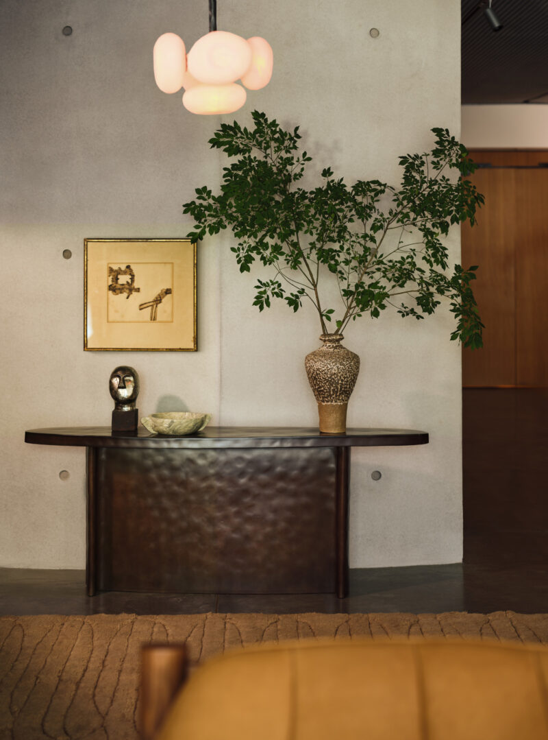 A dark, curved Swizz Beatz CB2 console table holds a metallic head sculpture, a stone bowl, and a tall vase with leafy branches against a concrete wall with a framed print and pendant light.