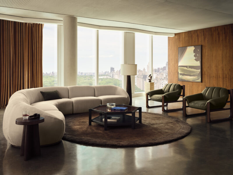 A modern Swizz Beatz CB2 living room with a curved sofa, two armchairs, a coffee table on a circular rug, and floor-to-ceiling windows overlooking a city skyline.