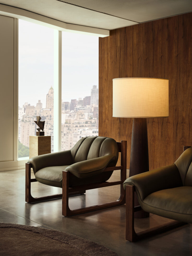 Two olive green Swizz Beatz CB2 leather armchairs and a tall floor lamp in a modern room with a city skyline view through large windows.
