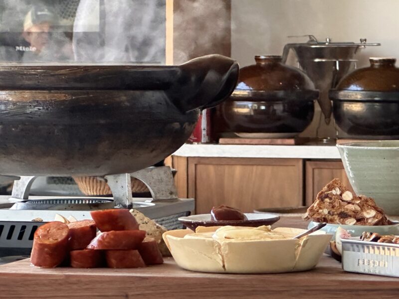 A steaming donabe pot on a burner next to a wooden board with sliced sausage, a small dish of spread, and crackers.