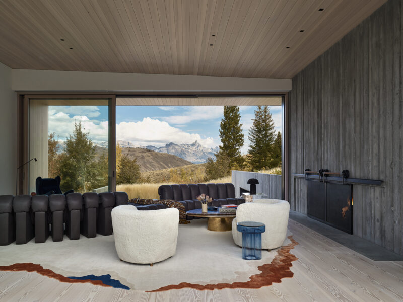 Living room with a dark sofa and two cream-colored armchairs facing a large window wall that overlooks a mountain landscape.