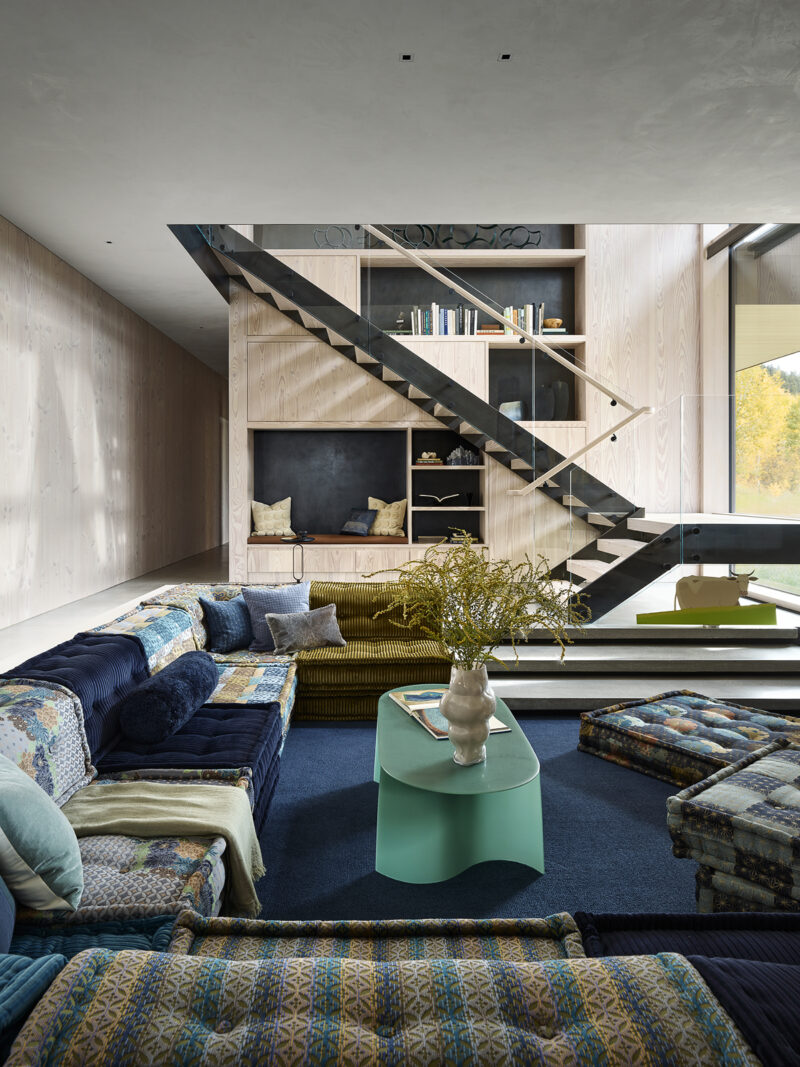 Living room with a large, multi-patterned sectional sofa, a green coffee table, and an open staircase leading to built-in wooden shelving.