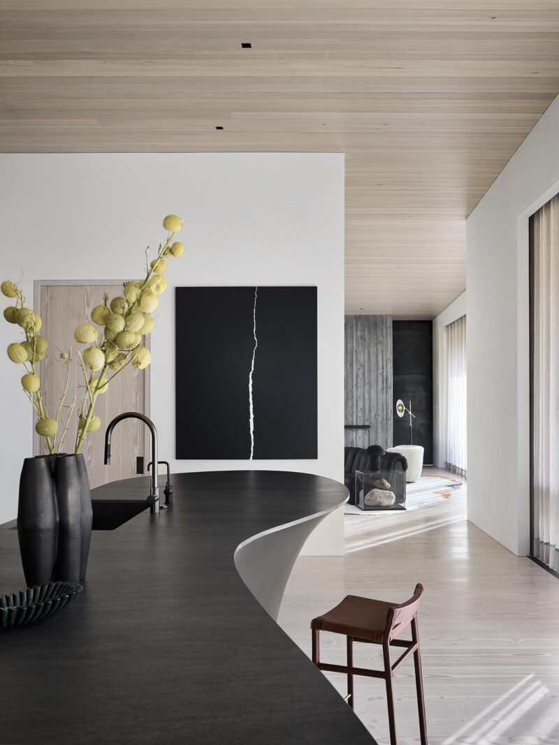 A modern kitchen island with a curved black countertop and a vase of yellow flowers, leading to an open-concept living area with a large abstract black painting.