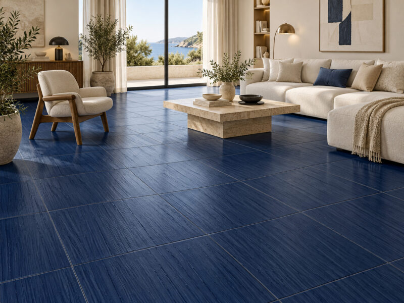 A living room with deep blue textured floor tiles, a beige sofa, a wood-frame armchair, and a view of the ocean through large glass doors.